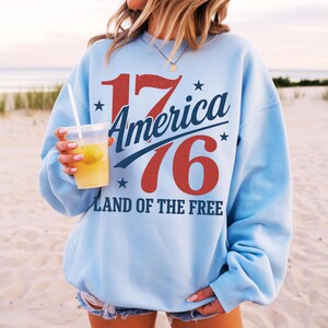 May include: Pale blue sweatshirt featuring the text "1776 America Land of the Free" in red and blue. The sweatshirt has a crew neck and long sleeves. The person in the image is holding a clear plastic cup with a yellow drink.