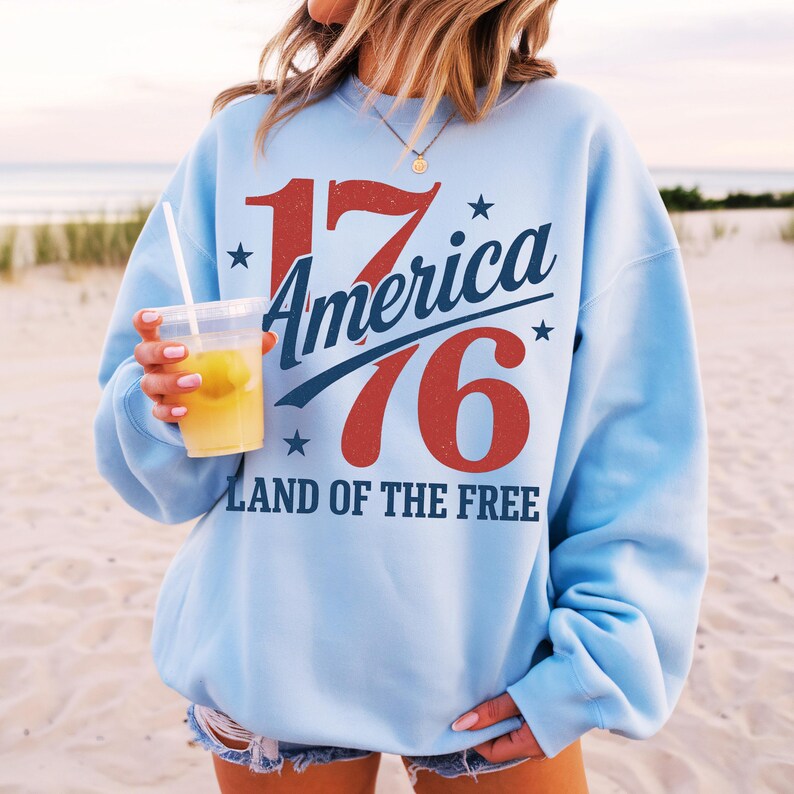 May include: Pale blue sweatshirt featuring the text "1776 America Land of the Free" in red and blue. The sweatshirt has a crew neck and long sleeves. The person in the image is holding a clear plastic cup with a yellow drink.