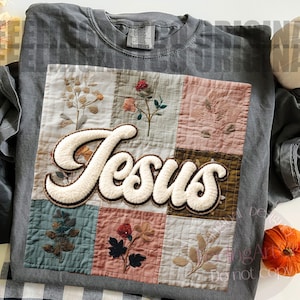 May include: A gray Comfort Colors t-shirt featuring a patchwork design with embroidered floral details. The word "Jesus" is prominently displayed in a fluffy, cream-colored font, adding a touch of texture and contrast to the design.