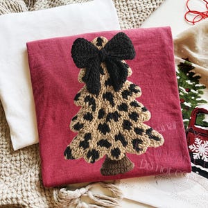 May include: A red decorative pillow with a knitted Christmas tree design. The tree features a leopard print pattern and a black bow at the top. A white pillow and a Christmas-themed towel are also visible.