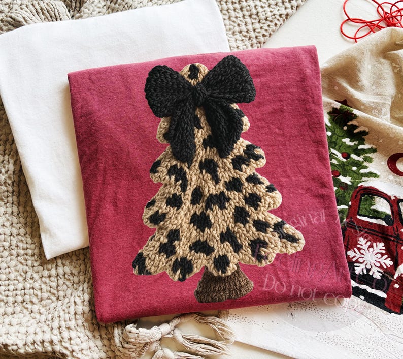 May include: A red decorative pillow with a knitted Christmas tree design. The tree features a leopard print pattern and a black bow at the top. A white pillow and a Christmas-themed towel are also visible.