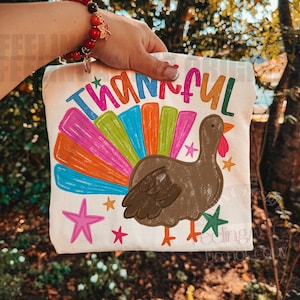 May include: A white t-shirt featuring a graphic of a brown turkey with a colorful, multi-striped tail in shades of pink, blue, orange, and green. The word Thankful is printed in a multi-colored font above the turkey, surrounded by small stars.