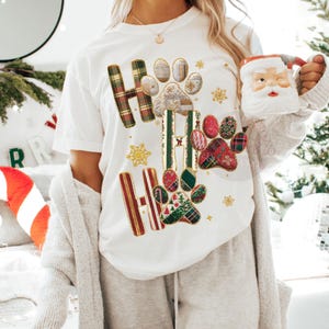 K&ouml;nnte beinhalten: Wei&szlig;es T-Shirt mit festlichem "Ho Ho Ho"-Design mit Pfotenabdr&uuml;cken und Schneeflocken. Die Buchstaben "H" sind mit Karomustern in Rot, Gr&uuml;n und Gold verziert. Eine Weihnachtsmann-Tasse wird gehalten.