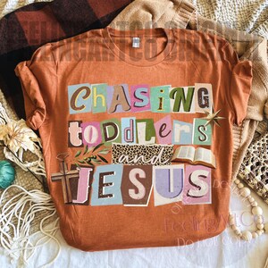 Pu&ograve; includere: T-shirt arancione con la scritta "Chasing Toddlers and Jesus" in un design colorato in stile collage. Il design include una croce, un libro aperto e una stella. La maglietta &egrave; realizzata in materiale morbido.