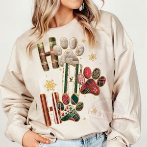 K&ouml;nnte beinhalten: Beiges Sweatshirt mit den Worten "Ho Ho Ho" in einem Patchwork-Design. Die Buchstaben "H" sind kariert und die Buchstaben "O" sind Pfotenabdr&uuml;cke mit verschiedenen Mustern. Goldene Schneeflocken und Sterne sind um das Design herum verstreut.