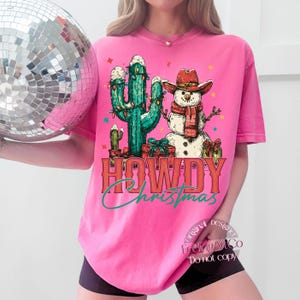 Peut inclure: Un t-shirt rose vif avec un motif de No&euml;l. Le design comprend un cactus, un bonhomme de neige portant un chapeau de cowboy et une &eacute;charpe, et les mots "HOWDY Christmas" dans une police de style western. Le t-shirt est de coupe ample.