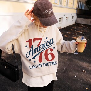 May include: A cream-coloured sweatshirt with the text "17 America 76 LAND OF THE FREE" in red, white, and blue. The image also shows a brown baseball cap with white text and a black purse.