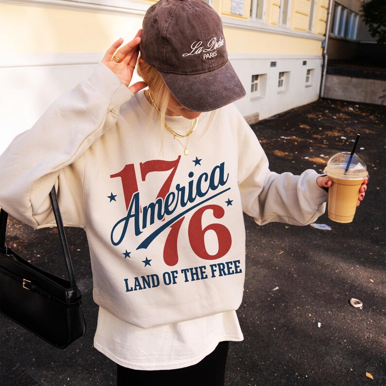 May include: A cream-coloured sweatshirt with the text "17 America 76 LAND OF THE FREE" in red, white, and blue. The image also shows a brown baseball cap with white text and a black purse.