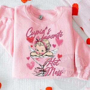 May include: Pink sweatshirt with a graphic of a baby cupid in a martini glass, surrounded by hearts. The text reads "Cupid's favorite Hot Mess" in pink script. The sweatshirt is folded.