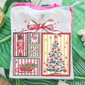 May include: White t-shirt with a Christmas-themed graphic. The design features a red bow, a nutcracker, Santa Claus, a star, a Christmas tree, and a sleigh filled with gifts. The shirt is folded with a pink and green shirt underneath.
