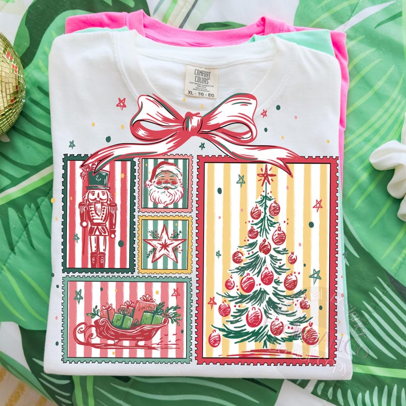 May include: White t-shirt with a Christmas-themed graphic. The design features a red bow, a nutcracker, Santa Claus, a star, a Christmas tree, and a sleigh filled with gifts. The shirt is folded with a pink and green shirt underneath.