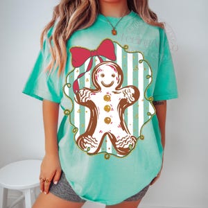 May include: A turquoise t-shirt featuring a gingerbread man design. The gingerbread man is brown with white icing and gold accents, set against a striped background with a red bow. The shirt has short sleeves.