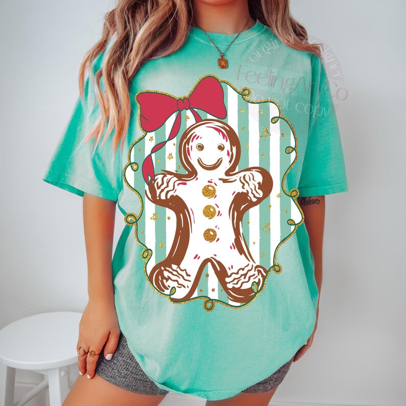 May include: A turquoise t-shirt featuring a gingerbread man design. The gingerbread man is brown with white icing and gold accents, set against a striped background with a red bow. The shirt has short sleeves.
