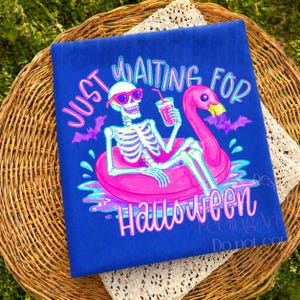 Peut inclure: Un t-shirt bleu avec un motif sur le th&egrave;me d'Halloween. Le design pr&eacute;sente un squelette portant des lunettes de soleil flottant sur un flotteur de flamant rose, tenant une boisson. Le texte dit "Just Waiting for Halloween."
