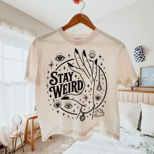 May include: Cream-colored t-shirt with a black graphic design. The design features a hand with long nails, eyes, stars, and the words "Stay Weird." The shirt is hanging on a wooden hanger.