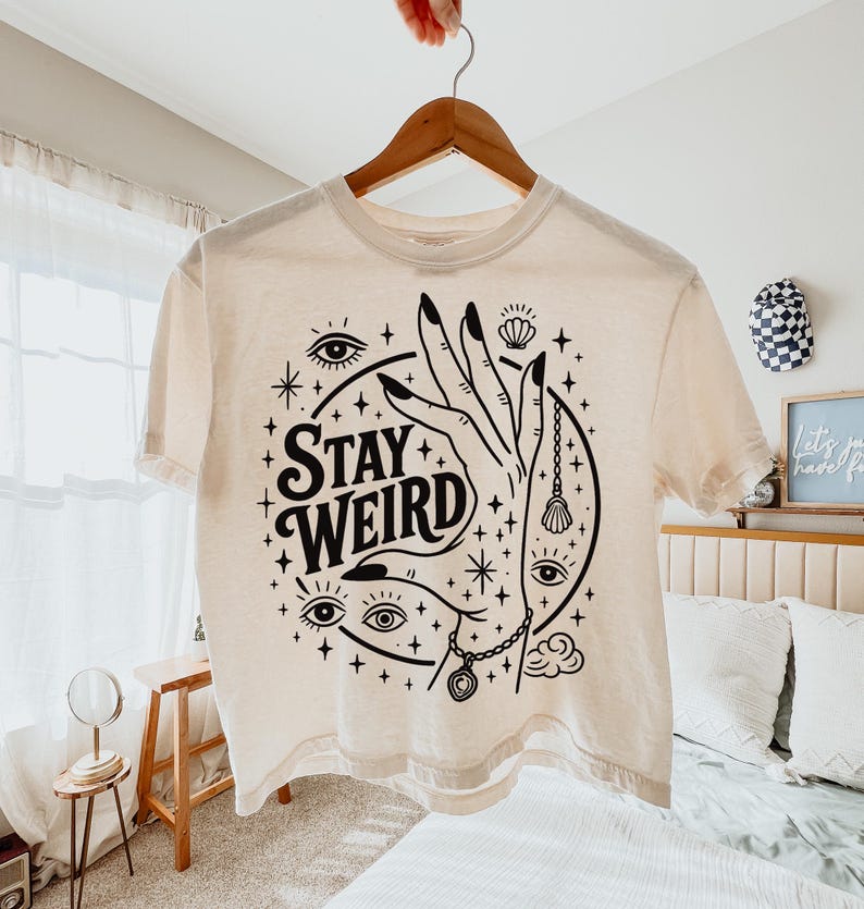 May include: Cream-colored t-shirt with a black graphic design. The design features a hand with long nails, eyes, stars, and the words "Stay Weird." The shirt is hanging on a wooden hanger.