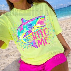 Op de afbeelding: Een neongeel t-shirt met een kleurrijke haai-afbeelding en de tekst "Bite Me". De haai is afgebeeld in tinten roze, blauw, groen en geel. Het shirt wordt gecombineerd met felroze shorts, wat een levendige outfit voor het strand cre&euml;ert.