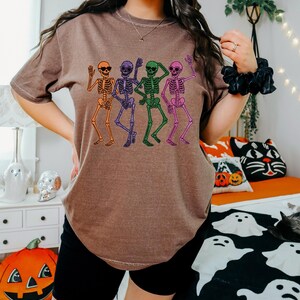 Puede incluir: Camiseta marr&oacute;n con un dise&ntilde;o de cuatro esqueletos bailando en naranja, morado, verde y rosa. Cada esqueleto lleva gafas de sol y levanta los brazos. Ideal para Halloween o eventos tem&aacute;ticos.