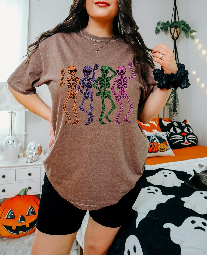 Puede incluir: Camiseta marr&oacute;n con un dise&ntilde;o de cuatro esqueletos bailando en naranja, morado, verde y rosa. Cada esqueleto lleva gafas de sol y levanta los brazos. Ideal para Halloween o eventos tem&aacute;ticos.