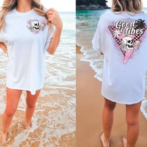 Puede incluir: Camiseta blanca de gran tama&ntilde;o con un gr&aacute;fico de calavera rosa y negro en la parte delantera y un gr&aacute;fico rosa y negro en la parte trasera que dice "Good Vibes" con una calavera y palmeras.