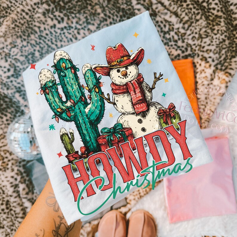 Peut inclure: T-shirt bleu clair avec un motif de No&euml;l. Le design pr&eacute;sente un bonhomme de neige portant un chapeau et une &eacute;charpe de cow-boy, un cactus orn&eacute; de guirlandes lumineuses et l'inscription "HOWDY Christmas" en rouge et vert.
