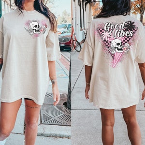 Puede incluir: Una camiseta beige de gran tama&ntilde;o con un gr&aacute;fico de calavera sosteniendo una bebida en la parte delantera y el texto "Good Vibes" con un gr&aacute;fico de calavera sosteniendo una bebida en la parte posterior.