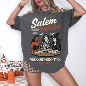May include: Dark gray t-shirt with the text "Salem Book Club" and an illustration of three witches reading books. The shirt also includes the text "Massachusetts Est. 1692" and "All Witches Welcome Meets Monthly".