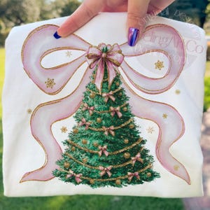 Pu&ograve; includere: T-shirt bianco con un design ad acquerello di un albero di Natale. L'albero &egrave; decorato con fiocchi rosa e ornamenti dorati, incorniciato da un grande fiocco rosa con accenti glitterati dorati. Fiocchi di neve dorati sono sparsi intorno al design.