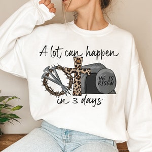 May include: White sweatshirt with a Christian Easter design. The design features a cross with a leopard print, a crown of thorns, nails, and a tomb with the text "He is Risen". The text "A lot can happen in 3 days" is written above the design.