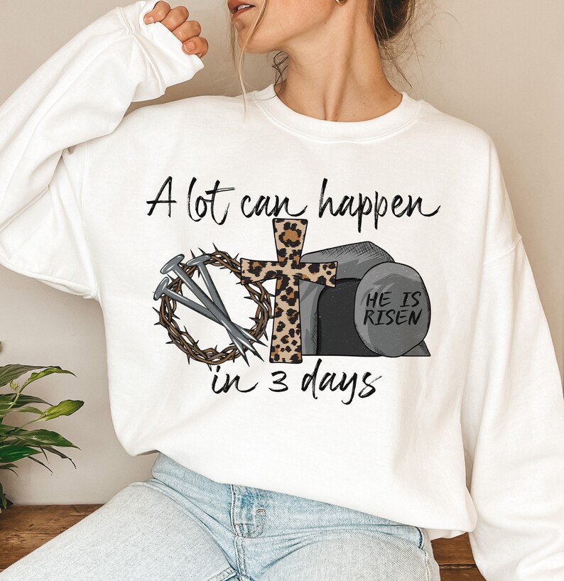 May include: White sweatshirt with a Christian Easter design. The design features a cross with a leopard print, a crown of thorns, nails, and a tomb with the text "He is Risen". The text "A lot can happen in 3 days" is written above the design.