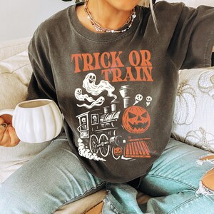 May include: Dark grey t-shirt with the words "TRICK OR TRAIN" in orange. The shirt features a Halloween-themed graphic of a train with ghosts, a jack-o'-lantern, and a skull. The person is holding a white pumpkin-shaped mug.