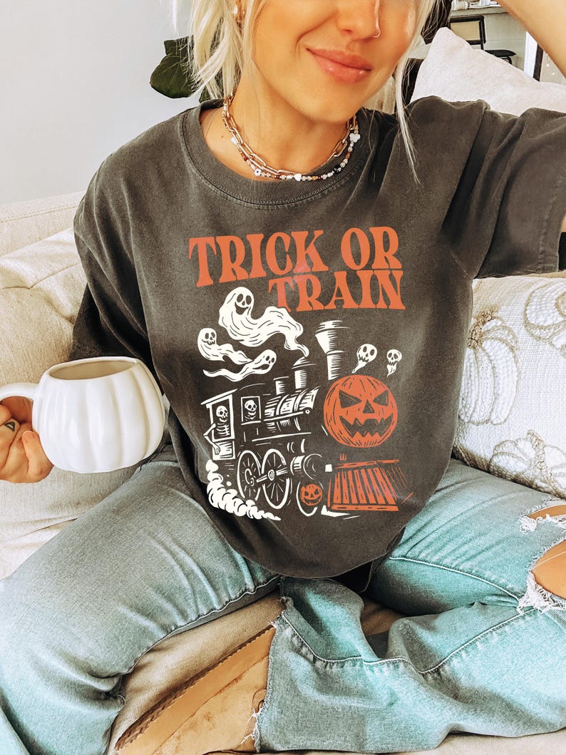 May include: Dark grey t-shirt with the words "TRICK OR TRAIN" in orange. The shirt features a Halloween-themed graphic of a train with ghosts, a jack-o'-lantern, and a skull. The person is holding a white pumpkin-shaped mug.