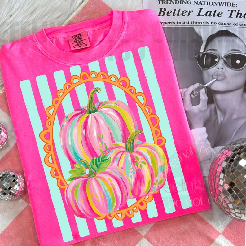 K&ouml;nnte beinhalten: Ein leuchtend pinkfarbenes Comfort Colors Sweatshirt mit einem bunten K&uuml;rbisdesign. Die K&uuml;rbisse sind mit rosa, gelben und blauen Streifen bemalt, vor einem hellblau gestreiften Hintergrund. Das Design wird von einem orangefarbenen, geschwungenen Rand umrahmt.
