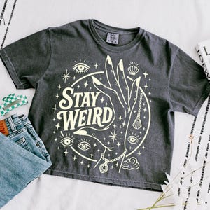 May include: Dark grey t-shirt with a cream-colored graphic featuring a hand, eyes, stars, and the words "Stay Weird." The shirt is paired with blue jeans and a green and white checkered hair clip.