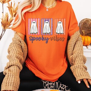May include: Orange t-shirt with a Halloween-themed design. The graphic features three white ghost figures with bows, set within rectangular frames. The words "spooky vibes" are written in a cursive font below the ghosts.