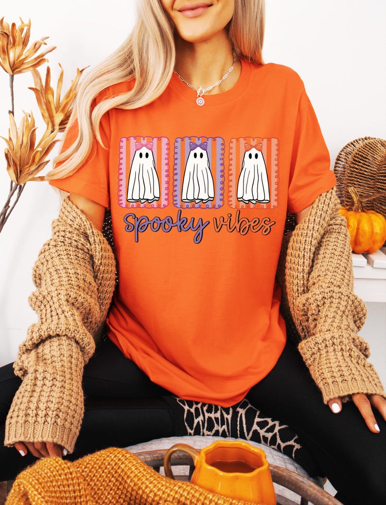 May include: Orange t-shirt with a Halloween-themed design. The graphic features three white ghost figures with bows, set within rectangular frames. The words "spooky vibes" are written in a cursive font below the ghosts.