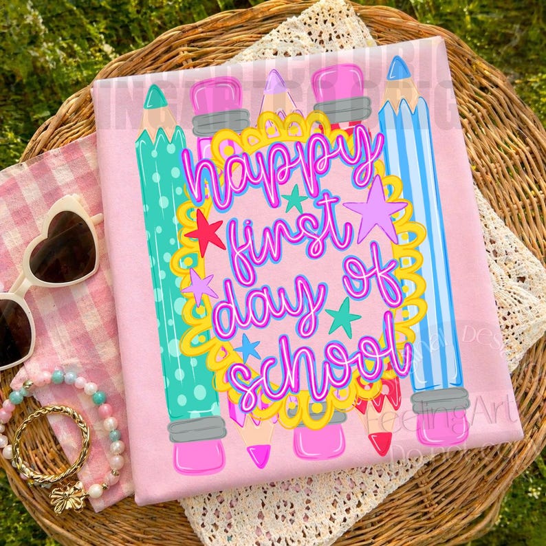 Puede incluir: Una camiseta rosa claro con un gr&aacute;fico de l&aacute;pices coloridos y un marco festoneado amarillo con el texto "happy first day of school" en letra cursiva rosa brillante. El dise&ntilde;o incluye peque&ntilde;as estrellas rosas y turquesas alrededor del texto.