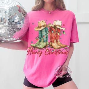 May include: A bright pink t-shirt features a graphic of two cowboy boots decorated with Christmas lights and cowboy hats. The text "Howdy Christmas" is written in a cursive font. A disco ball is in the background.