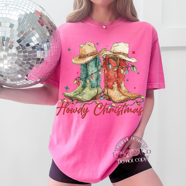 May include: A bright pink t-shirt features a graphic of two cowboy boots decorated with Christmas lights and cowboy hats. The text "Howdy Christmas" is written in a cursive font. A disco ball is in the background.