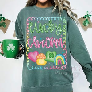 K&ouml;nnte beinhalten: Gr&uuml;nes Sweatshirt mit dem Schriftzug "Lucky Charm" in einem farbenfrohen, skurrilen Design. Das Design umfasst ein Herz, einen Goldtopf, ein Kleeblatt und einen Regenbogen. Eine gr&uuml;ne Tasse mit einem Kleeblatt wird in der linken Hand gehalten.