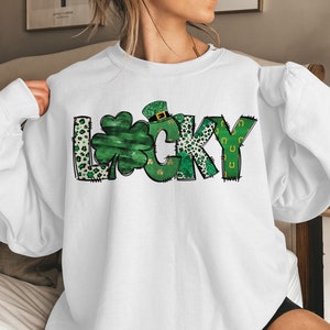 May include: White sweatshirt with the word "LUCKY" in green, black, and white. The letter "L" has a leopard print pattern, the "U" is a shamrock, and the "C" has a leprechaun hat. The "Y" has green horseshoes.