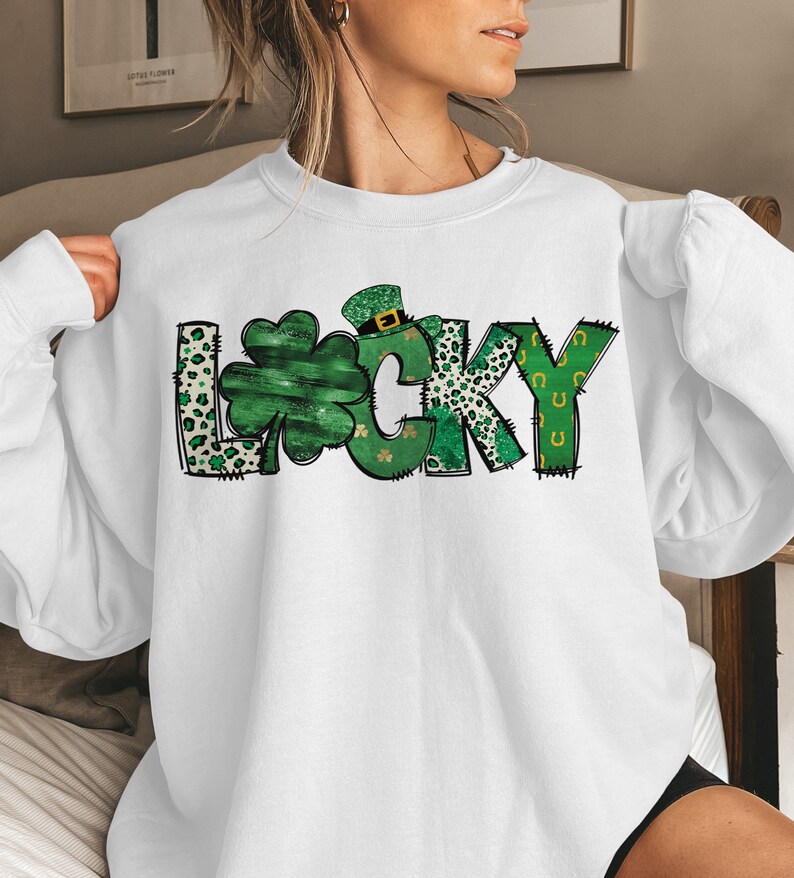 May include: White sweatshirt with the word "LUCKY" in green, black, and white. The letter "L" has a leopard print pattern, the "U" is a shamrock, and the "C" has a leprechaun hat. The "Y" has green horseshoes.