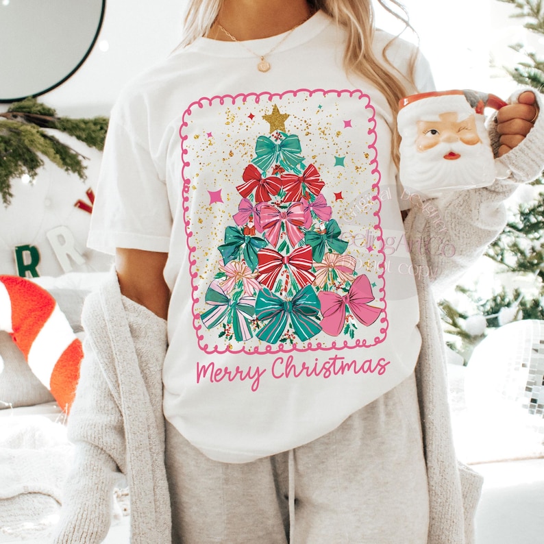 May include: White t-shirt with a Christmas tree design made of bows in red, pink, and teal, topped with a gold star. The words "Merry Christmas" are written in pink script below the tree.