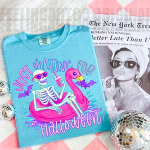 Peut inclure: Un t-shirt bleu clair avec l'inscription "Just Waiting For Halloween". Le graphisme repr&eacute;sente un squelette avec des lunettes de soleil, un flotteur flamant rose et des chauves-souris violettes. Le t-shirt est de la marque Comfort Colors.
