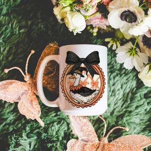 Peut inclure: Mug en c&eacute;ramique blanche avec un dessin de canard de dessin anim&eacute; portant un chapeau de sorci&egrave;re et un n&oelig;ud noir. Le canard est dans un cadre ovale orange avec un bord festonn&eacute;. La tasse a une grande anse et est entour&eacute;e de papillons et de fleurs d&eacute;coratifs.