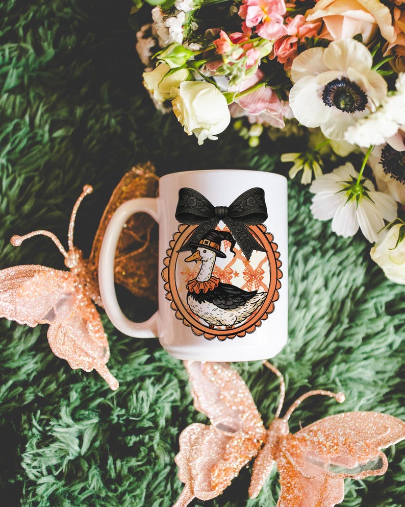 Peut inclure: Mug en c&eacute;ramique blanche avec un dessin de canard de dessin anim&eacute; portant un chapeau de sorci&egrave;re et un n&oelig;ud noir. Le canard est dans un cadre ovale orange avec un bord festonn&eacute;. La tasse a une grande anse et est entour&eacute;e de papillons et de fleurs d&eacute;coratifs.