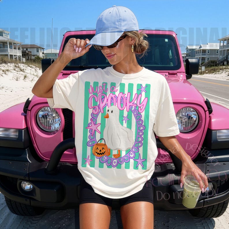 May include: A cream-colored short-sleeve t-shirt featuring a graphic of a white ghost-like duck holding a jack-o'-lantern, surrounded by a purple oval frame, green vertical stripes, and pink stars, with the text "just a SPOOKY" printed above.