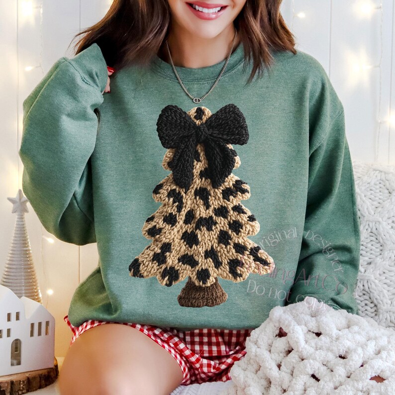 May include: A green sweatshirt featuring a knitted Christmas tree design with a black bow at the top. The tree has a leopard print pattern in tan and black. The sweatshirt is a crew neck style.