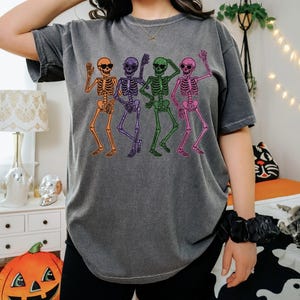 Puede incluir: Camiseta gris oscuro con un dise&ntilde;o de cuatro esqueletos bailando en naranja, morado, verde y rosa. Cada esqueleto lleva gafas de sol y saluda. La camiseta tiene un corte holgado y es ideal para un look casual.