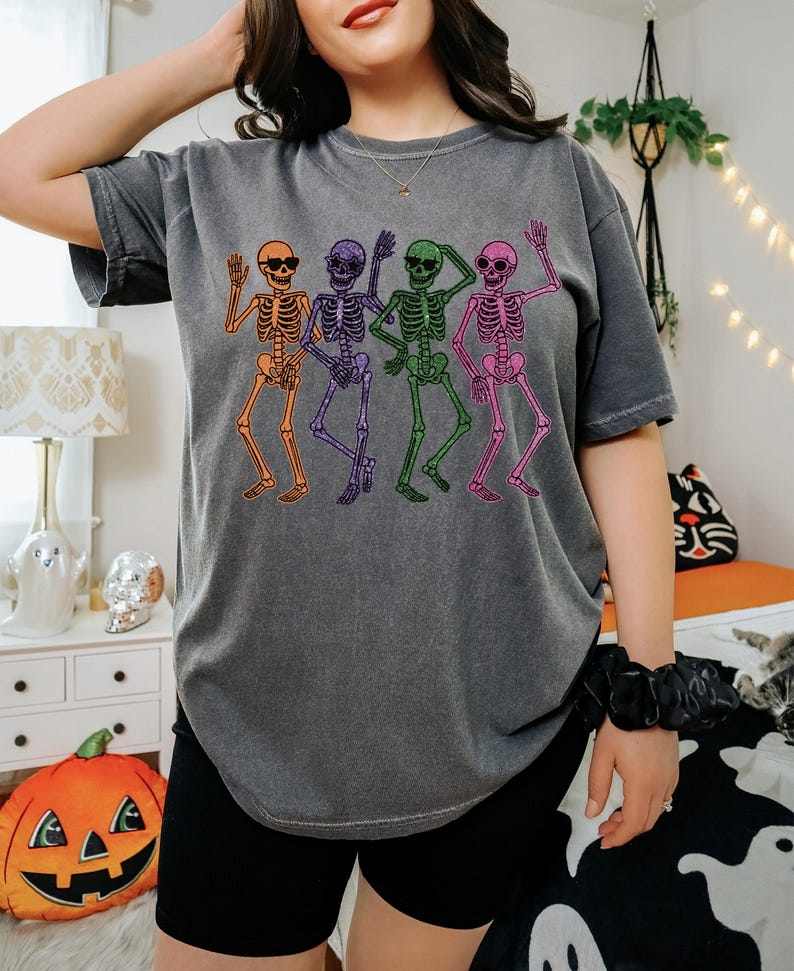 Puede incluir: Camiseta gris oscuro con un dise&ntilde;o de cuatro esqueletos bailando en naranja, morado, verde y rosa. Cada esqueleto lleva gafas de sol y saluda. La camiseta tiene un corte holgado y es ideal para un look casual.
